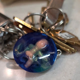 Keychain with a blue green creepy cute baby with bum showing trapped in resin charm on a light-colored surface