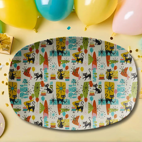 An oval party platter with a colorful retro 1950s pattern featuring black cats, surfboards, stars, and birthday candles on a cream background. The platter is styled on a light yellow surface with balloons, confetti, and a gift box in the background.
