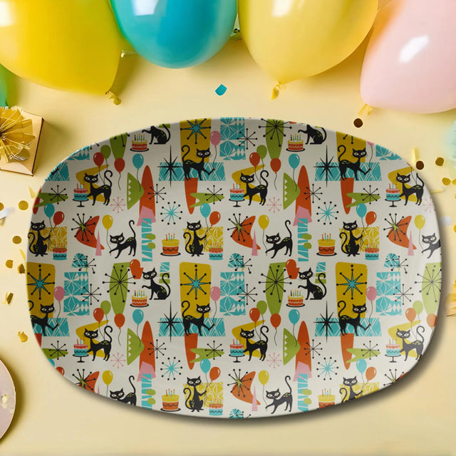 An oval party platter with a colorful retro 1950s pattern featuring black cats, surfboards, stars, and birthday candles on a cream background. The platter is styled on a light yellow surface with balloons, confetti, and a gift box in the background.