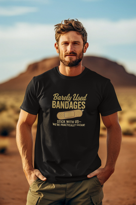 A man standing in a desert setting, wearing a black T-shirt with a retro graphic that says 'Barely Used Bandages'.
