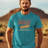 A man standing in a desert setting, wearing a blue T-shirt with a retro graphic that says 'Barely Used Bandages'.