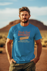 A man standing in a desert setting, wearing a blue T-shirt with a retro graphic that says 'Barely Used Bandages'.