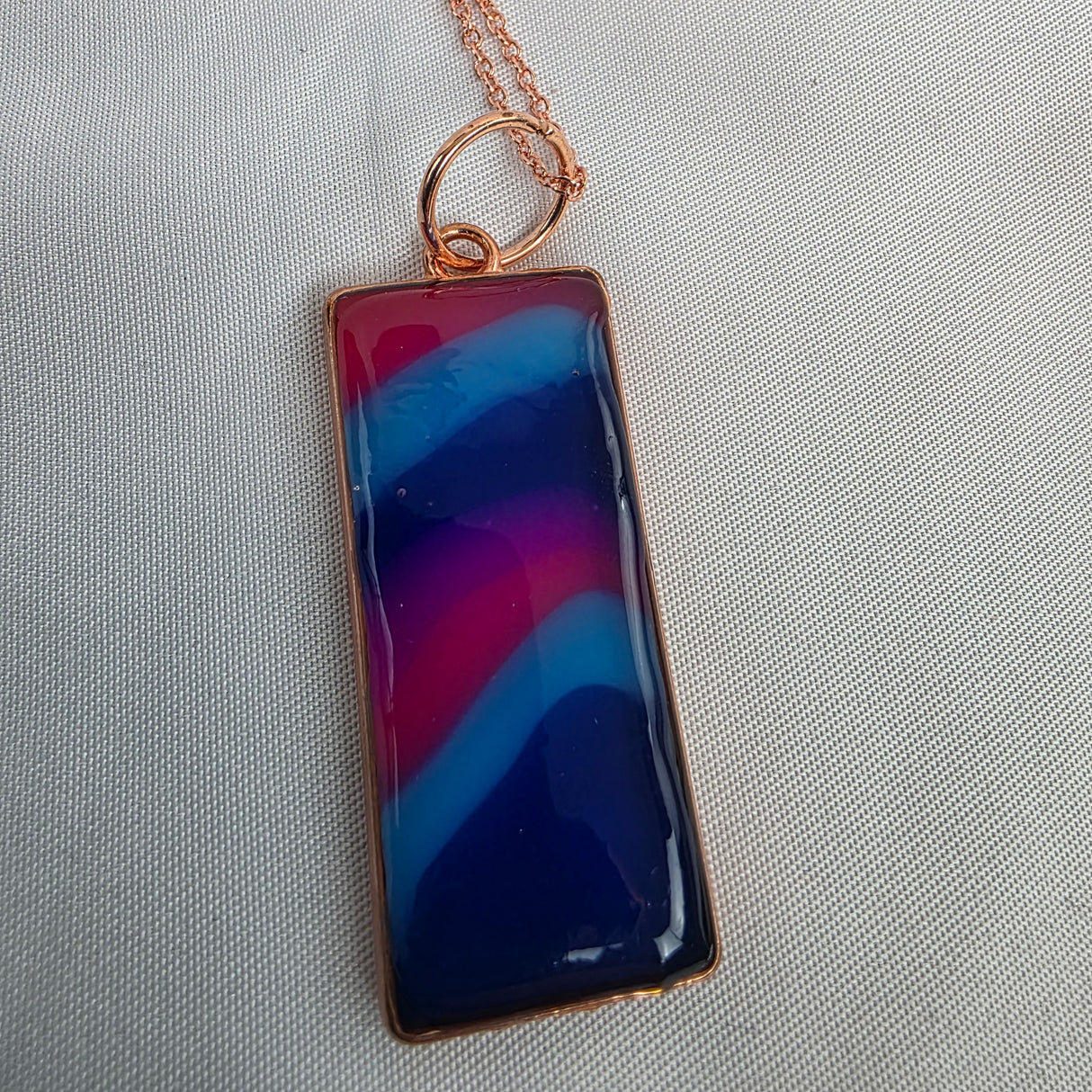 Rectangular pendant with blue and red design on a gold chain against a light gray background