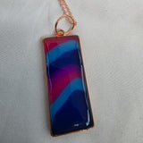 Rectangular pendant with blue and red design on a gold chain against a light gray background